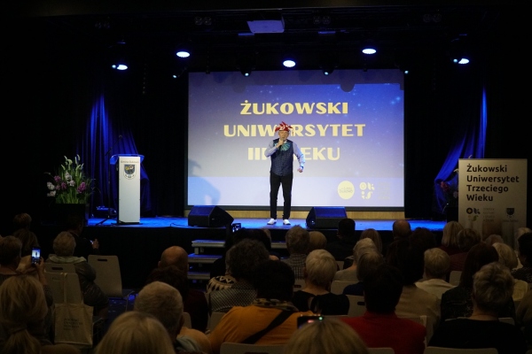 na scenie Andrzej Rosiewicz, w tle napis Żukowski Uniwersytet Trzeciego Wieku, przed sceną zebrani studenci i studentki.