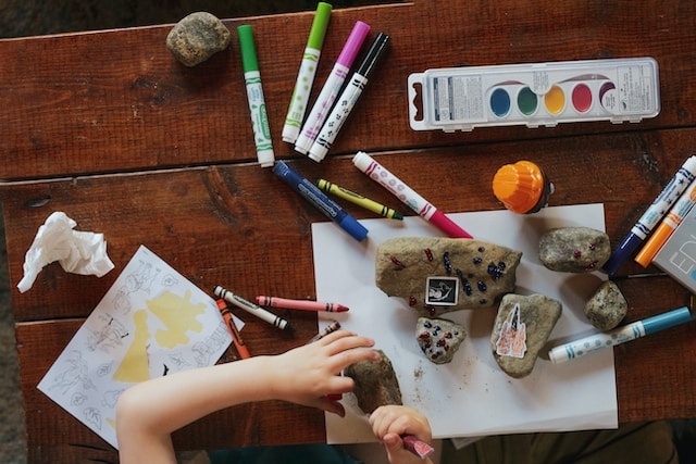 Na biurku leżą pisaki, farbki, kartki, kamienie. Dziecko trzyma w dłoniach kamień i go maluje.