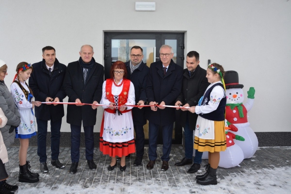 Na zdjęciu pośrodku mężczyzn stoi kobieta, po obu stronach stoją dzieci i trzymają wstążkę, a mężczyźni i kobieta przecinają wstążkę. 