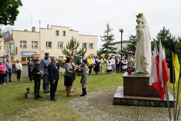 na pierwszym planie kobiety, za nimi stoją mężczyźni. kobieta wręcza dziecku wiązankę kwiatów. po prawej stronie pomnik Maryi. w tle koniety i mężczyźni zgromadzeni wokół pomnika.